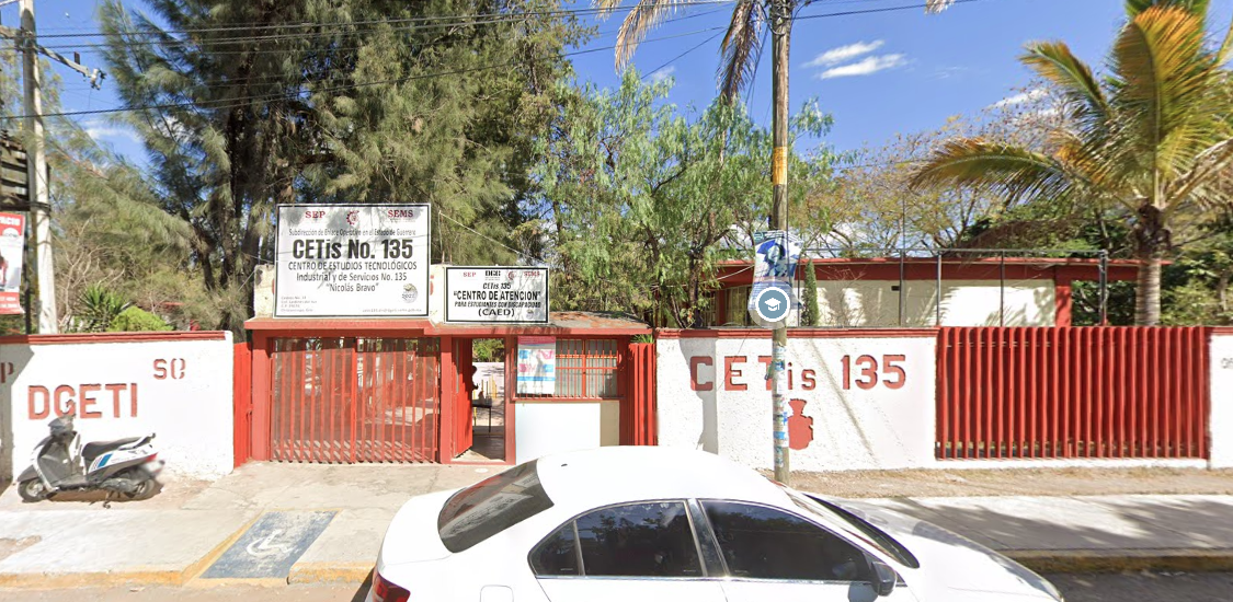 Fotografía en la que se muestra la entrada del plantel, conformada por una reja de color rojo que da acceso a las instalaciones. Sobre la rej<a hay una placa con los datos del plantel. Las instalaciones están rodeadas por muros blancos intercalados con rejas rojas y sobre uno de los muros está escrito en letras rojas CETIS 135.